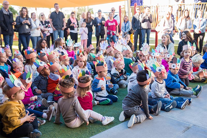 A group of young children wearing handmade paper hats with colorful feathers sit on the ground during an outdoor event, with adults standing in the background.